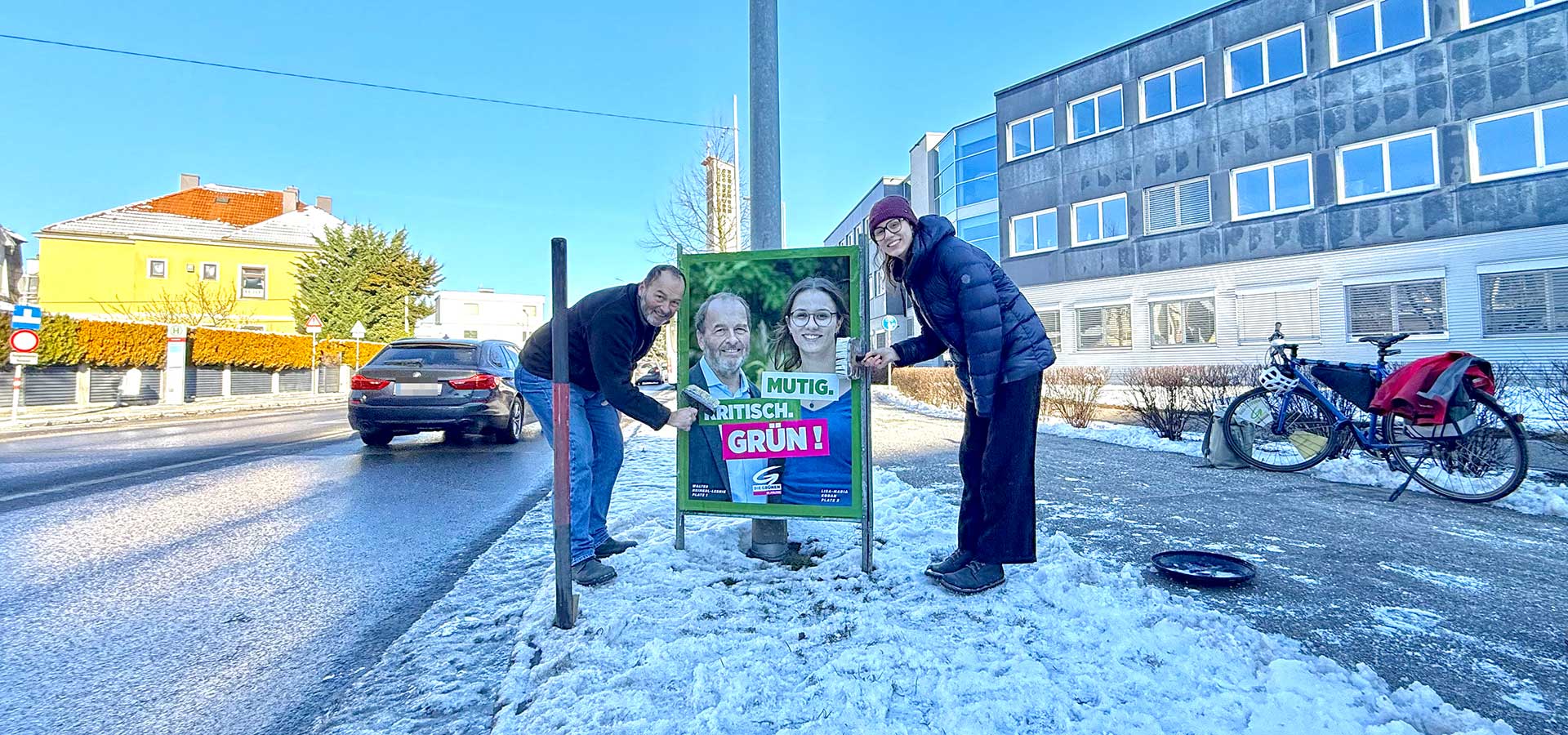 Walter Heimerl-Lesnik und Lisa-Maria Koban affichieren ein Wahlkampfplakat am Grünstreifen entlang einer Straße