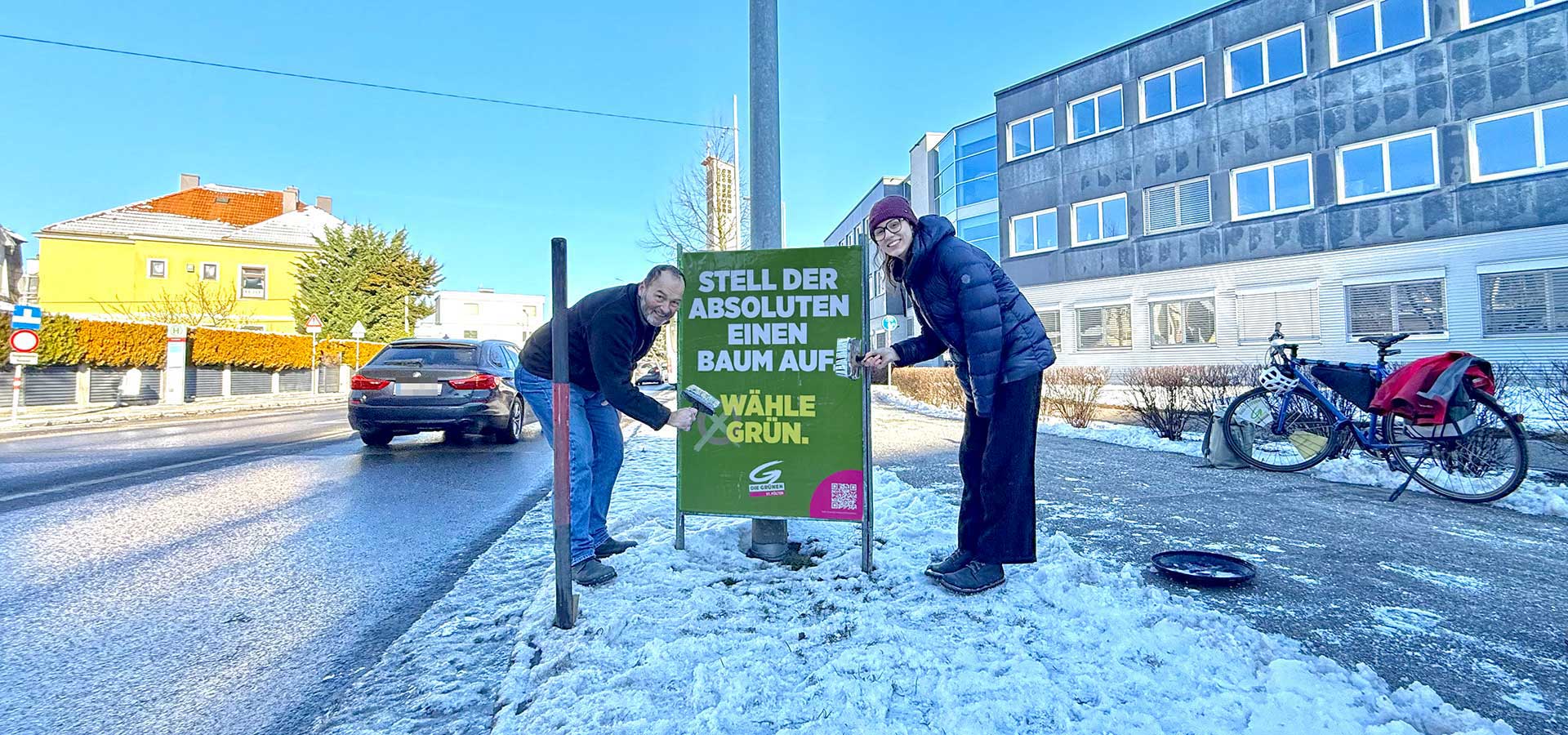 Walter Heimerl-Lesnik und Lisa-Maria Koban affichieren ein Wahlkampfplakat am Grünstreifen entlang einer Straße