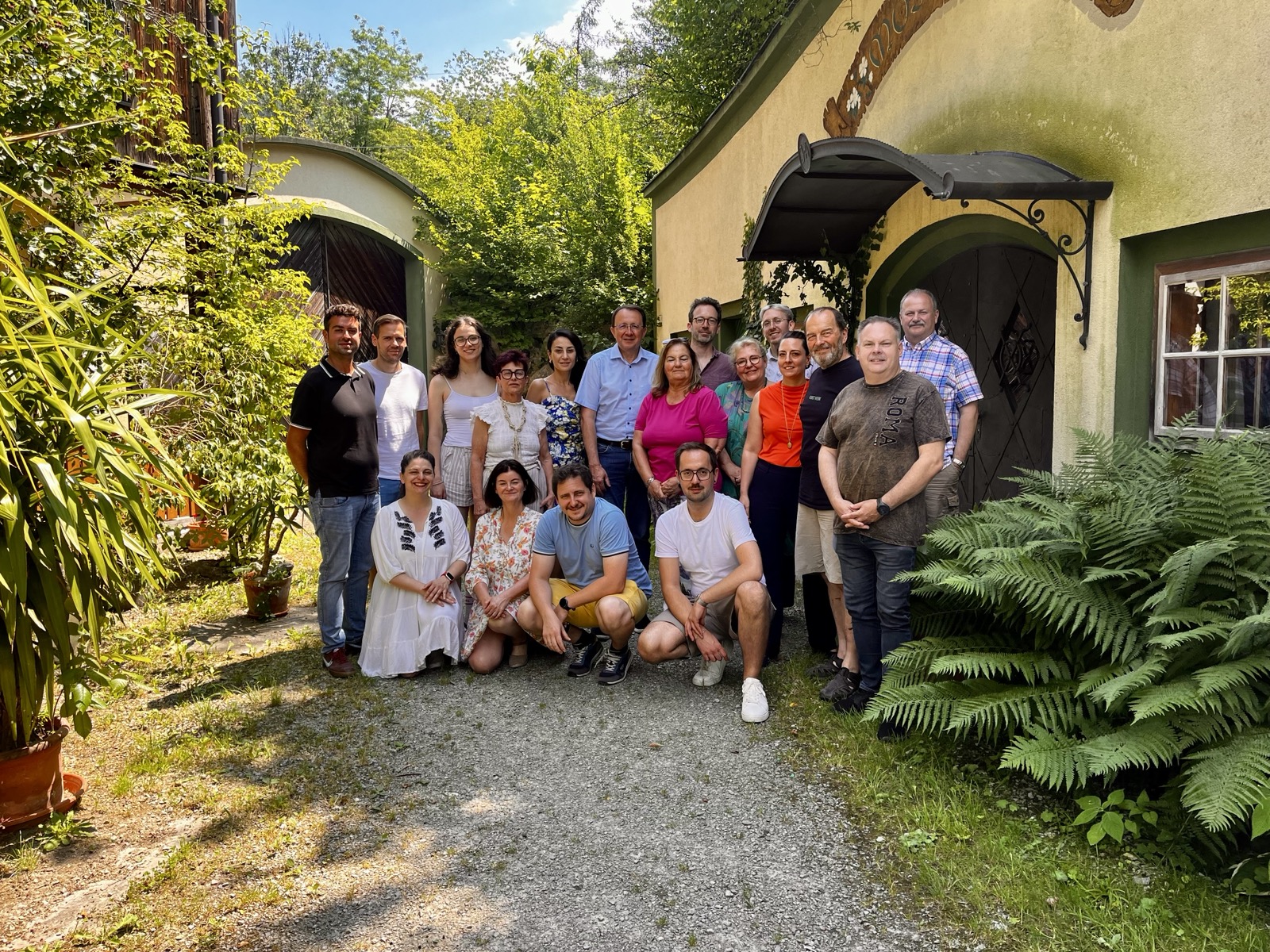 SPÖ- und Grüne Gemeinderät*innen stehen und sitzen in einem sonnigen Innenhof vor begrünten Gebäuden. Die Gruppe ist im Rahmen einer gemeinsamen Klausur versammelt.