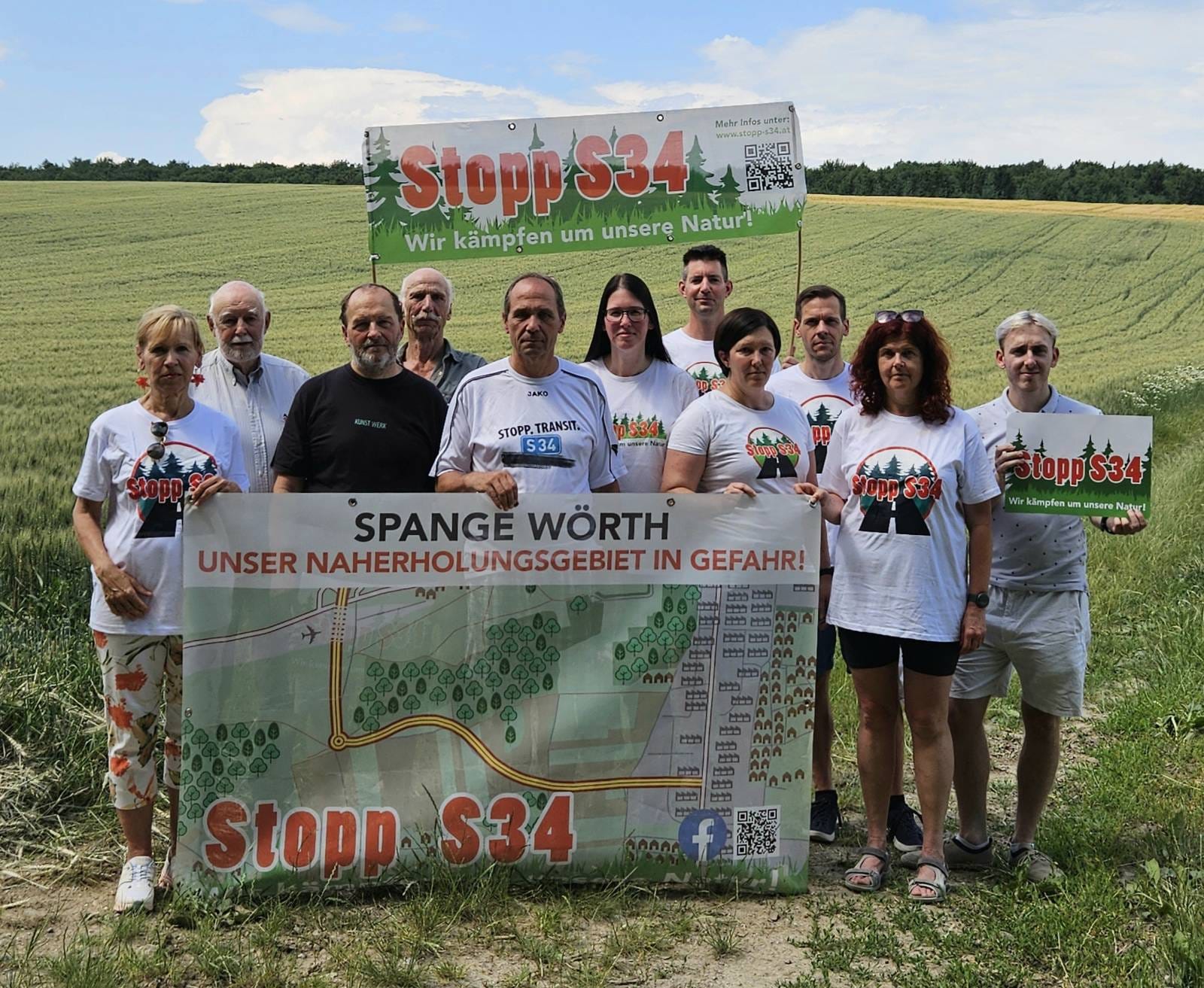 Eine Gruppe von mehreren Personen steht vor einem Feld hinter einem Transparent mit der Aufschrift „Spange Wörth – Unser Naherholungsgebiet in Gefahr!“ sowie einem Banner „Stopp S34 – Wir kämpfen um unsere Natur!“. Die Personen setzen sich gegen das Straßenbauprojekt ein.