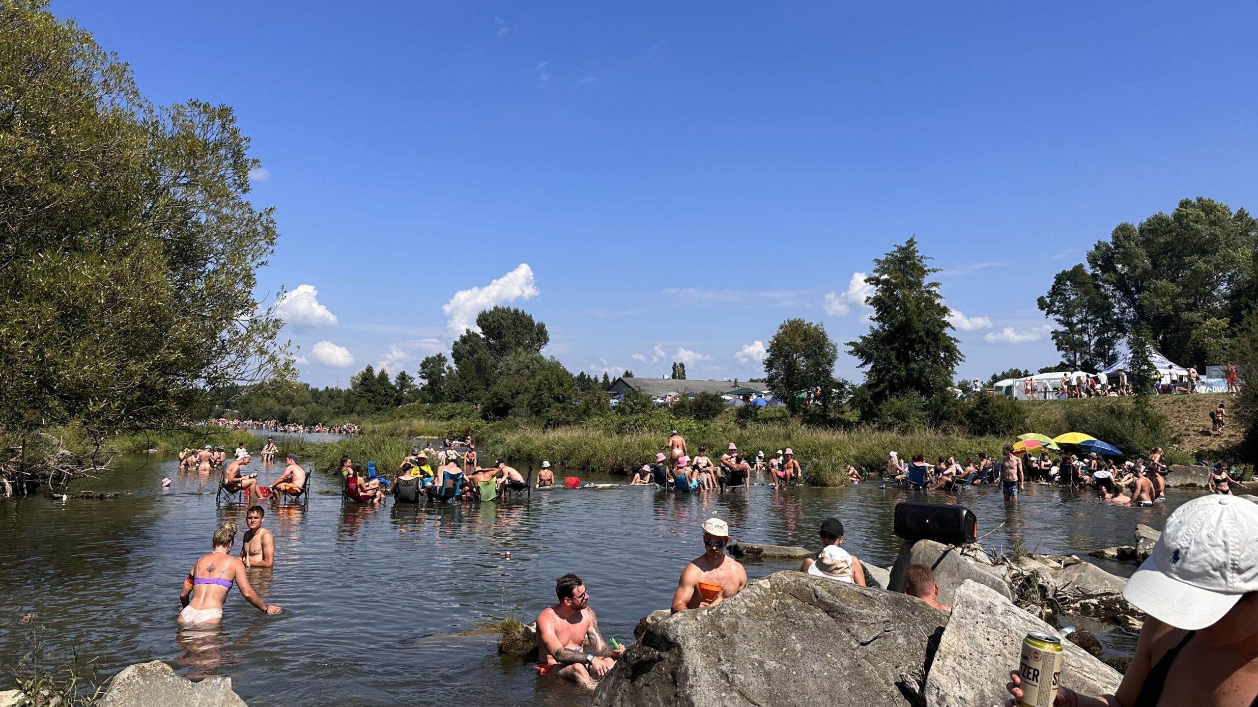 Viele Menschen baden und sitzen im flachen Wasser der Traisen an einem sonnigen Sommertag. Entlang des Flussufers halten sich zahlreiche weitere Besucher:innen auf, im Hintergrund sind Bäume, Wiesen und Festivalzelte zu sehen.