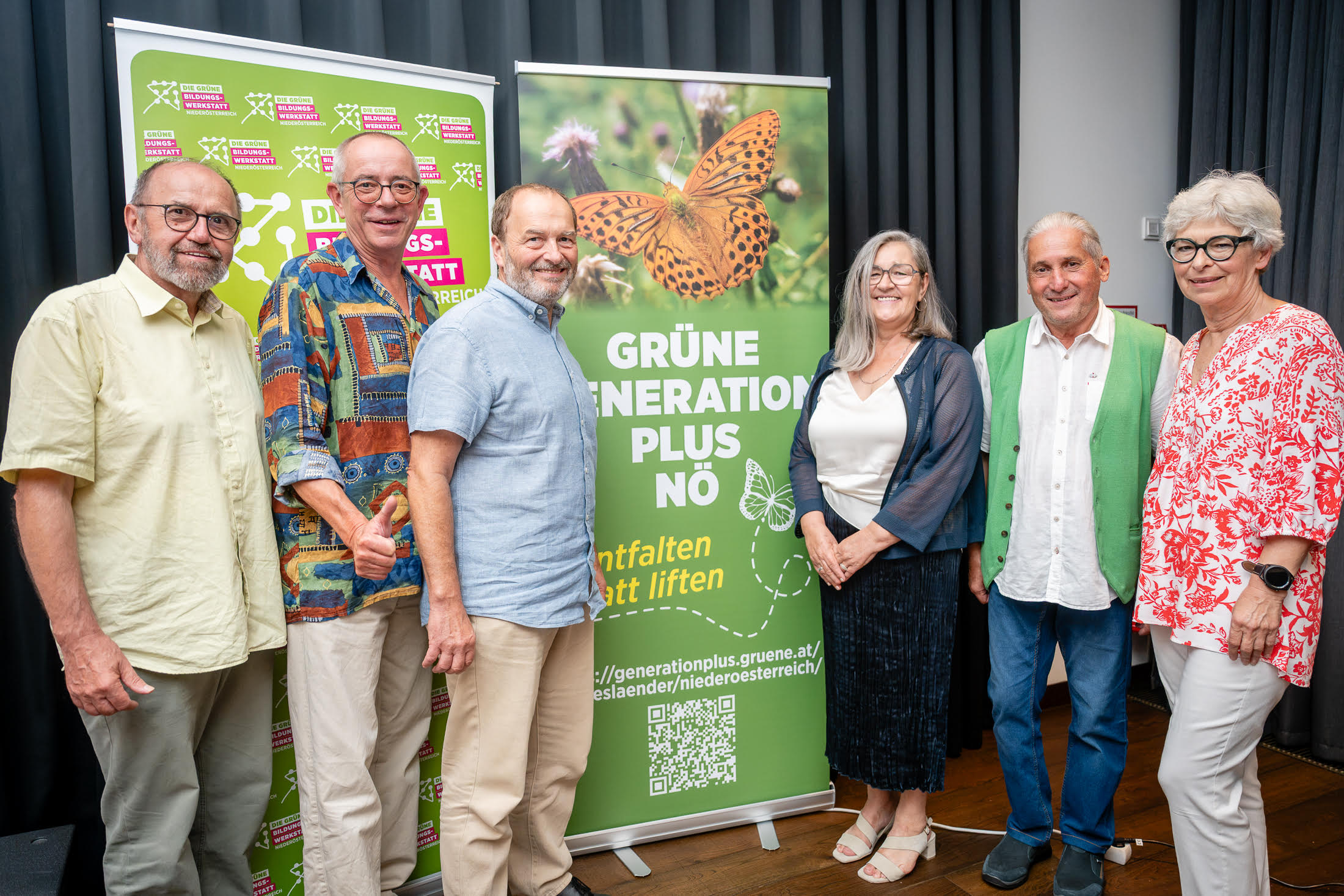 Sechs Personen stehen lächelnd nebeneinander in einem Innenraum vor dunklen Vorhängen und zwei Roll-up-Bannern der Grünen Parteiorganisation.